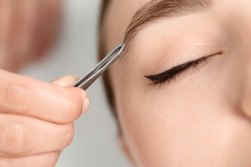 Beautician plucking young woman's eyebrow on light background, closeup