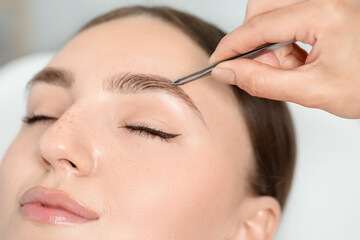 Beautician plucking young woman's eyebrow in beauty salon, closeup