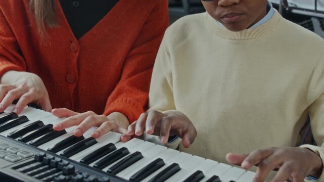 Tilt down shot of young female intern and African American little school boy playing on digital piano together during music class