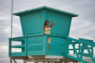 Pretty young woman with green eyes in a yellow silk suit and pink heels, hand over her eyes looking at the sea, leaning on a railing of a green lifeguard hut. Concept beauty, beach, coast, ocean.