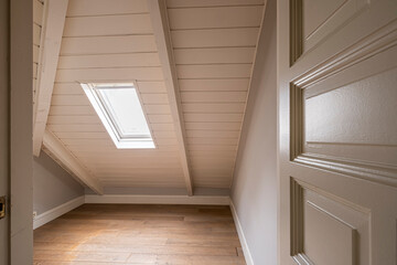 The photograph captures an empty attic room featuring a slanted roof and a small skylight window,...