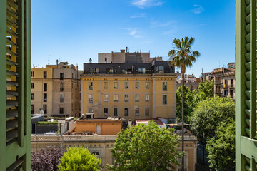 Fototapeta premium A scenic daytime view of urban buildings surrounded by greenery is seen through open green shutters, emphasizing architecture and nature's coexistence in Barcelona Spain