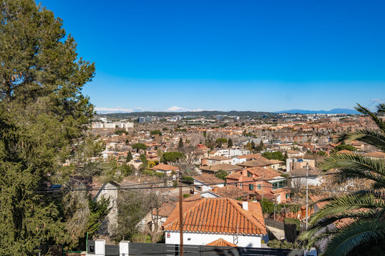The image captures a scenic view of a cityscape from an elevated point, showcasing red brick rooftops, green foliage, and a clear blue sky stretching into the horizon Sant Cugat in Spain