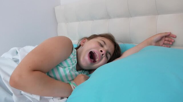 A little tired brunette girl with loose hair sleeps in a crib at home, tossing from side to side and yawning with her mouth wide open. White blanket, blue pillow.