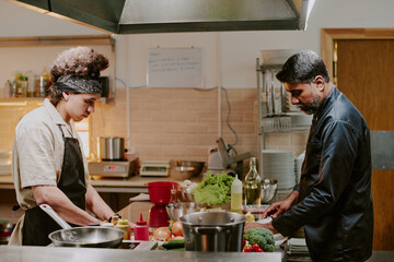 Middle aged chef dressed in black uniform and his young sous chef cooking dishes together in modern restaurant kitchen