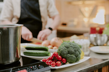 Medium close up of white ceramic plate full of fresh tomatoes and broccoli on steel kitchen counter, cook cutting vegetables in blurred background