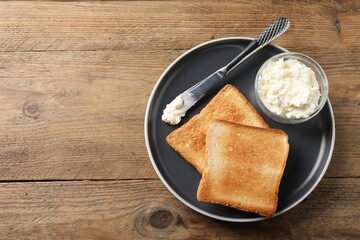 Delicious toasted bread slices with cream cheese and knife on wooden table, top view. Space for text