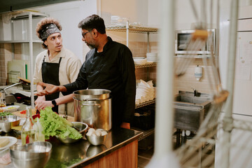 Chef and his sous chef discussing extra ingredients of recipe while sous chef chopping tomatoes