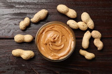 Tasty peanut butter in bowl and nuts on wooden table, flat lay