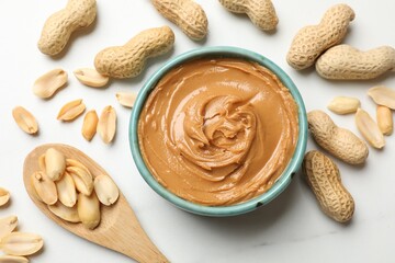 Tasty peanut butter in bowl and spoon with groundnuts on white table, flat lay