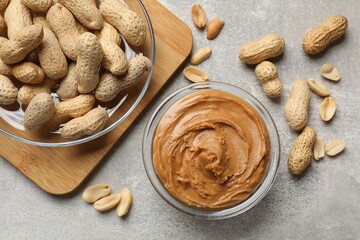 Tasty peanut butter in bowl and groundnuts on grey textured table, flat lay