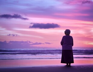 Fototapeta premium Silhouette of an old woman on the beach staring at the dreamy pastel violet pink colorful sky of sunset or sunrise