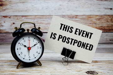 This event is postponed text on paper card with alarm clock on wooden background