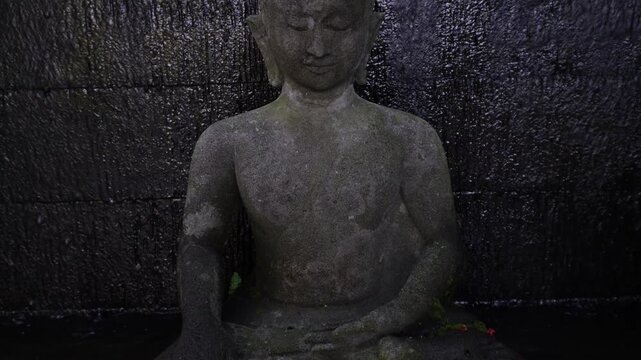 Stone statue of a Buddha figure meditating facing the front with waterfall on stone wall background. Bodhisattva Face. Concept for Vesak Day and Enlightenment Day. Empty blank copy text space