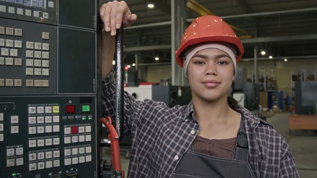 Chest up slowmo portrait of young Latin workwoman in safety overalls posing by industrial control panel and looking confidently at camera, fixing orange hard hat on head in manufacturing factory