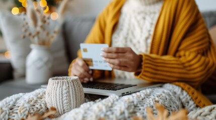 A person, wrapped in a cozy blanket, holds a credit card while shopping online on a laptop, embodying the comfort of autumn and the convenience of modern technology.