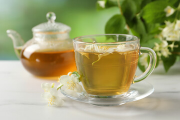Aromatic jasmine tea in cup, flowers, teapot and green leaves on white marble table