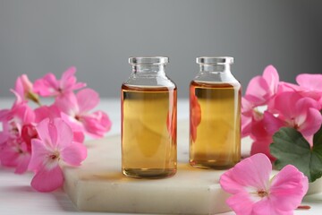 Bottles of geranium essential oil and beautiful flowers on white table, closeup