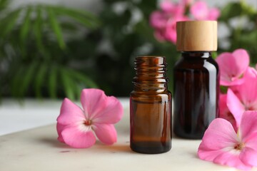 Bottle of geranium essential oil and beautiful flowers on white table, closeup