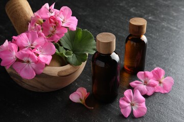 Bottles of geranium essential oil, beautiful flowers and mortar with pestle on black table