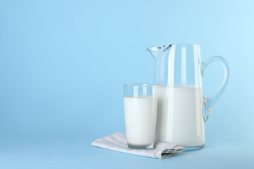 Jug and glass of fresh milk on light blue background, space for text