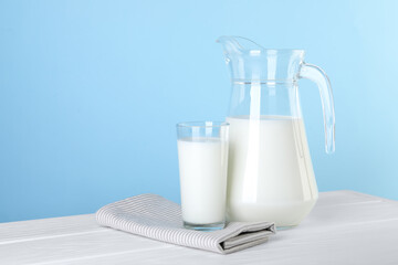 Jug and glass of fresh milk on white wooden table against light blue background, space for text