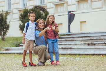 Fototapeta premium A smiling Caucasian woman kneels beside her son and daughter, all dressed for daytime, as she happily escorts them to school, with a campus building in the background.