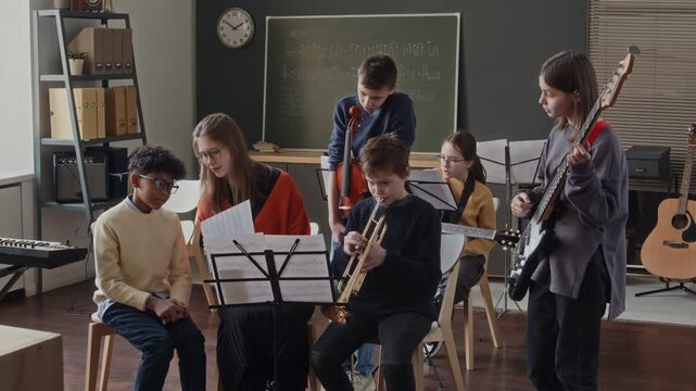 Young female intern sitting in classroom surrounded by four school children while teaching them to read sheet music, girl examining notes in background