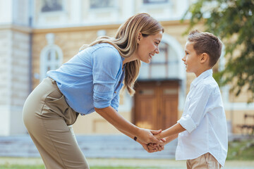 Obraz premium A smiling woman and her young son engage in a loving exchange by a school, reflecting a moment of daily family life with their casual attire and backpacks.