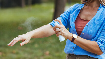 A Caucasian woman dressed casually applies mosquito spray to her arm outdoors, ensuring skin...