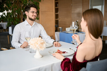 Happy young couple sitting in the restaurant while holding glasses with white wine on dating