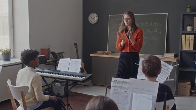 Young school teacher making final conducting gesture and applauding group of little school kids after successful performance in music class