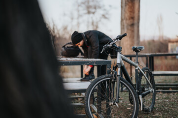 A young man in casual attire takes a break, resting his head on a park bench, beside his mountain...