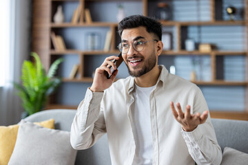 Indian young man in glasses and casual clothes is sitting on the sofa at home and smilingly talking...