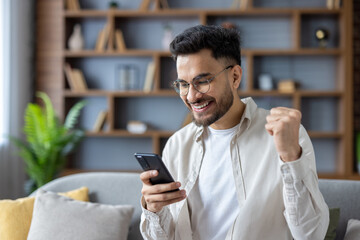 Close-up photo of a young Indian man wearing glasses at home, sitting on the sofa, looking at the...