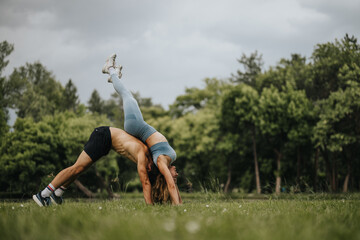 Fitness couple performing acro yoga poses outdoors in a park setting. Energetic and healthy lifestyle in nature surrounded by trees.