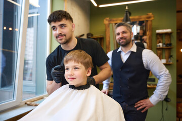 Little satisfied boy watching stylish barber combing his hair