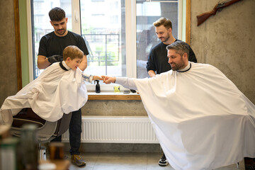 Little boy giving fist bump to father who sitting in barber chair
