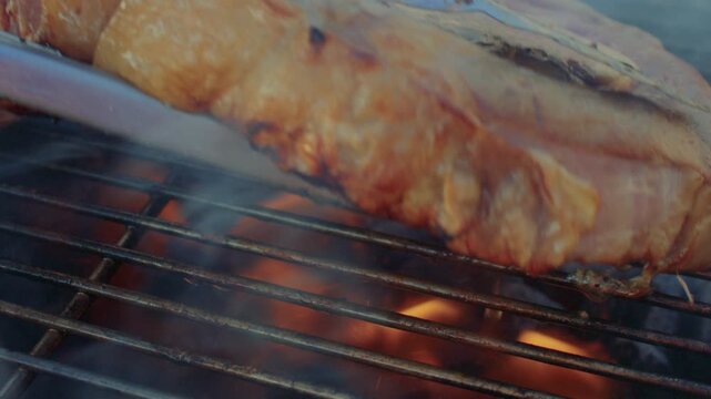 Close up shot of kitchen tongs taking and flipping a half-cooked beef steak on a roasting grate. Cook flips a ribeye steak on barbeque brazier, revealing the grilled side.