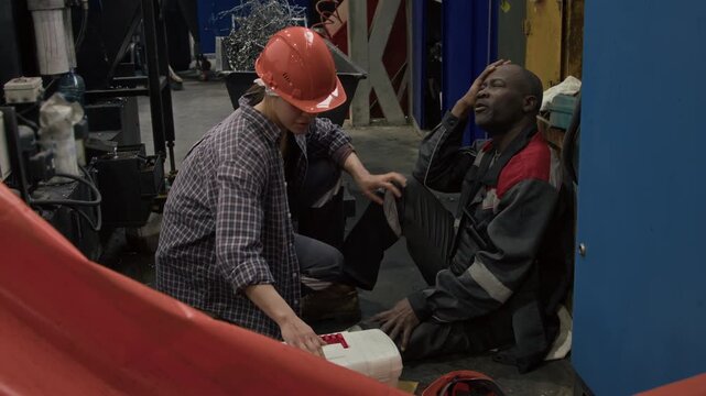 Side full shot of workwoman in hardhat with first aid kit running towards workman with work-related head injury, administering first aid at factory workshop