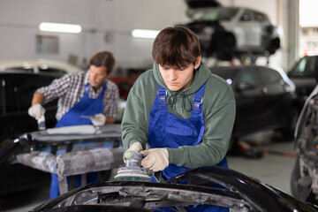 Young auto technician sanding car part using power sander with concentration, highlighting professional vehicle maintenance and repair skills in workshop