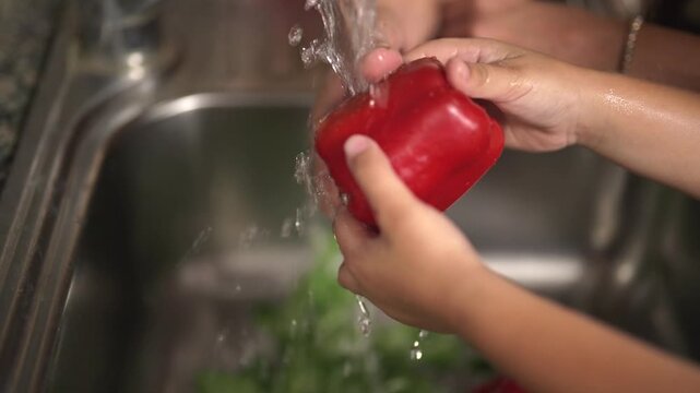 Manos de ni&ntilde;o y su madre lavando pimiento rojo en la cocina para el almuerzo en c&aacute;mara lenta