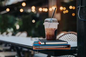 Close-up of an iced coffee on top of notebooks at an outdoor cafe table with warm bokeh lights in the background.