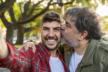 A happily married gay couple is smiling and kissing while taking selfies.