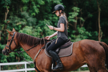 Young girl riding a horse on a ranch while using a smart phone, showcasing multitasking and...
