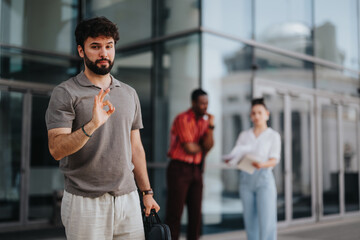 Confident man making an okay gesture while his colleagues stand in the background outside a modern business building, suggesting teamwork and positivity.