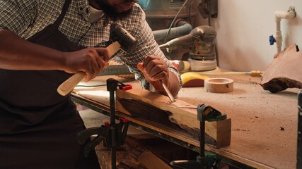 African american manufacturer shaping raw timber using chisel and hammer, creating wood art, making...
