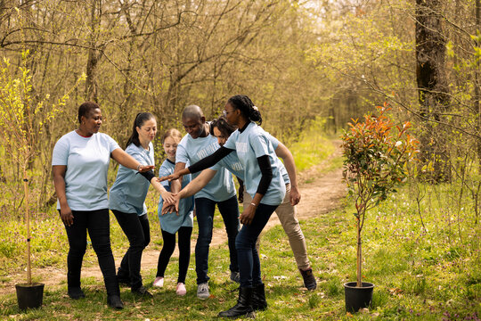 Cheerful enthusiastic team of activists unite to plant trees in the woods, taking action to protect natural environment. People celebrating their voluntary work by connecting hands together.