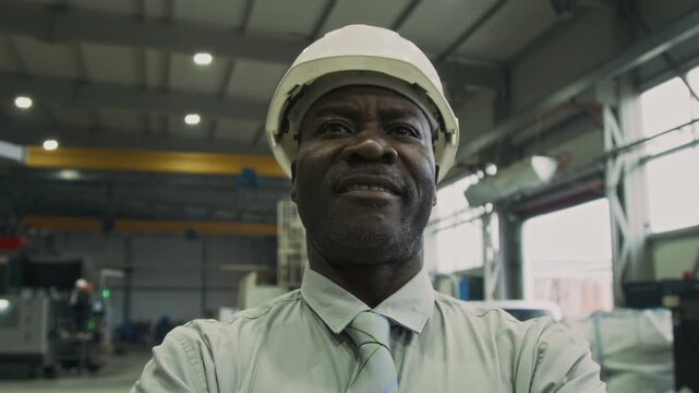 Tilt low angle shot of confident African American businessman in white hardhat, shirt and tie crossing arms and hero posing in manufacturing workshop, looking straight and smiling
