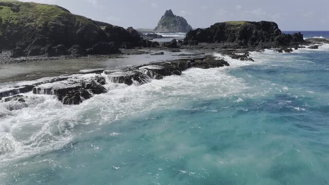 Drone flies over blue waters toward Buraco da Raquel surrounded by shallow pools at low tide and Ilha Sela Gineta looming ahead in Fernando de Noronha, Pernambuco, Brazil
